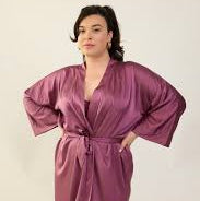 A woman modeling a shiny purple satin robe with wide sleeves and a tie waist, set against a plain light background.