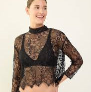 A woman modeling a sheer black floral lace mock-neck long-sleeve top over a black bralette, paired with black underwear, against a plain off-white studio background.