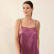 A woman modeling a short satin slip dress in a dusty mauve-purple color, standing against a plain off-white background.