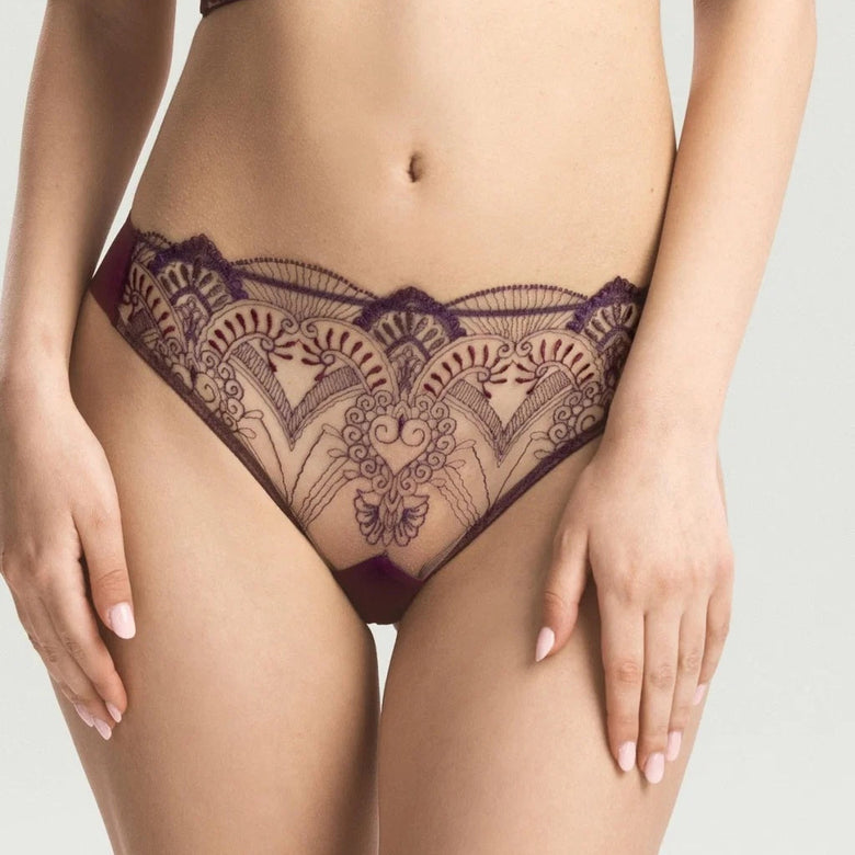A model wearing a dark purple lace lingerie set, featuring a sheer bralette with floral lace detailing and matching high-cut lace panties, shown against a light gray studio background.