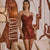A woman models a short, burgundy satin slip dress with lace trim in a modern living room setting with a beige sofa and a decorative floor lamp.
