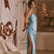 A woman models a light blue satin slip dress with a thigh-high slit in a modern living room setting with a beige sofa and abstract wall art.