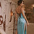 A woman wearing a light blue satin slip dress with a high thigh slit and lace-up back detail, standing in a styled indoor room with a sofa and wooden table.