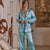 A woman wearing a light blue satin pajama set with black piping stands in a modern living room setting. The set includes a long-sleeve button-front top with a tie waist and matching wide-leg pants.