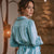 A woman wearing a light blue satin pajama set with a matching belt, shown from the back in an indoor setting with warm lighting.