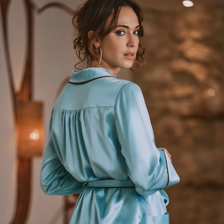 A woman wearing a light blue satin pajama set with a matching belt, shown from the back in an indoor setting with warm lighting.