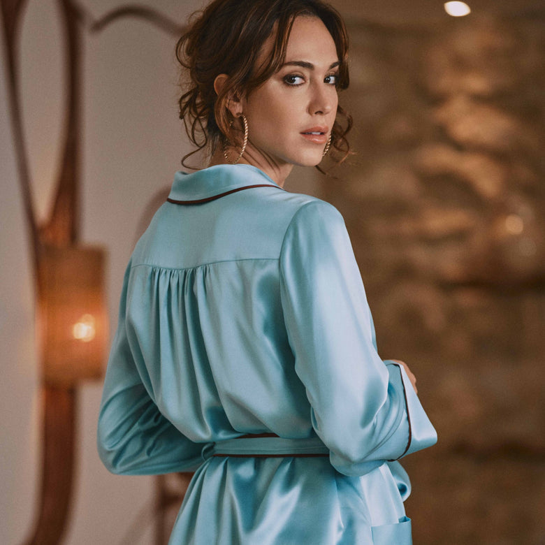 A woman wearing a light blue satin pajama set with a matching belt, shown from the back in an indoor setting with warm lighting.