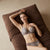 A woman reclines on a brown cushion wearing a matching lace bra and underwear set in a warm neutral-toned indoor setting.