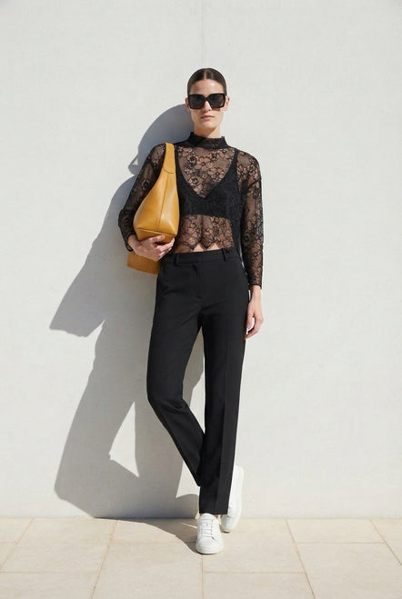 A model stands against a light gray wall wearing a sheer black lace mock-neck top, black trousers, white sneakers, and sunglasses, while carrying a tan shoulder bag.