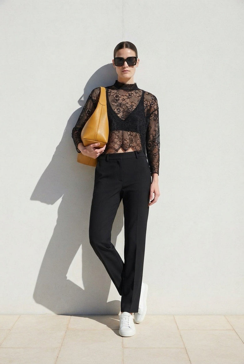 A model stands against a light gray wall wearing a sheer black lace mock-neck top, black trousers, white sneakers, and sunglasses, while carrying a tan shoulder bag.