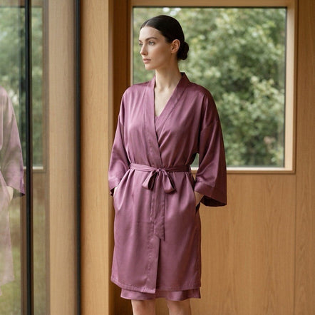 A woman wearing a mauve-purple silk-like robe stands barefoot in a modern room with wood paneling and a window looking out onto green trees.